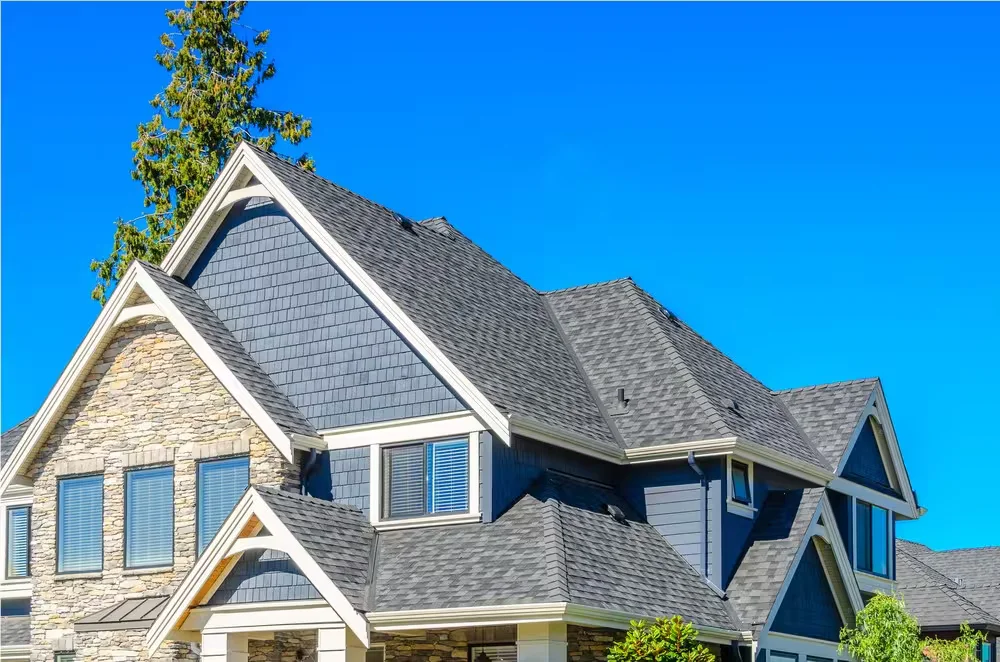 Two-story house with a gray shingle roof, blue shingle and lap siding, and a stone-clad gable under a clear blue sky