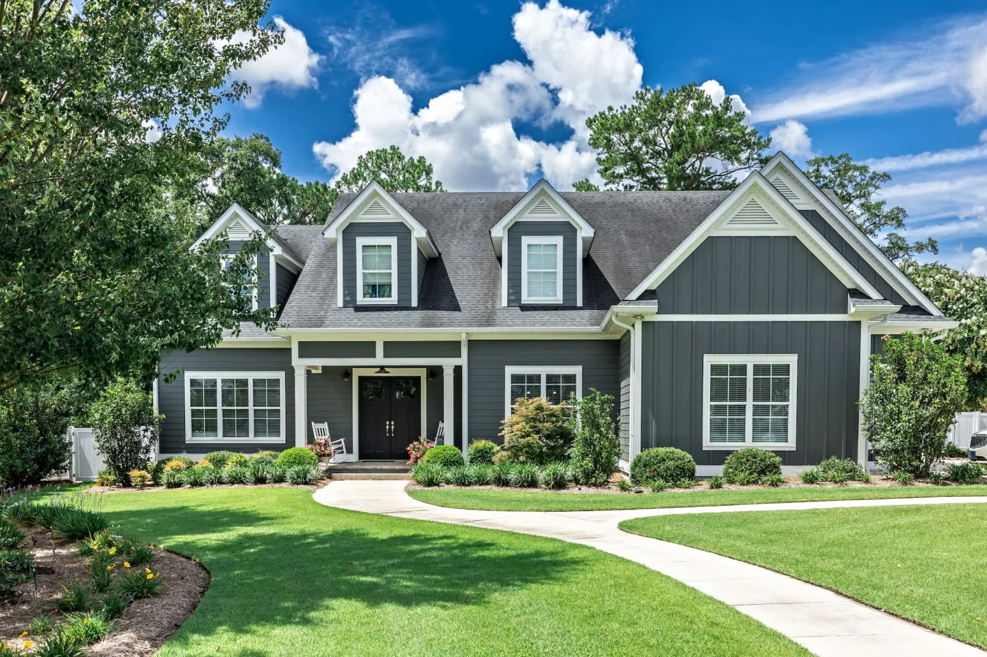 Gray two-story house with white trim, dormers and a black double entry door behind a green lawn and curved walkway