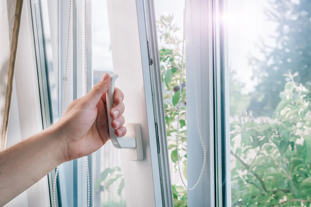 Hand opening a white window sash by its handle, with green trees outside
